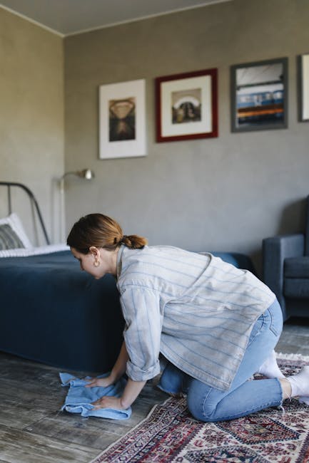 A woman with brown hair tied back, dressed in a light striped shirt and blue jeans, is kneeling on a patterned area rug in a modern bedroom. She is using a blue cloth to clean the wooden floor next to a dark grey bed frame. The room has beige walls decorated with three framed pictures, a grey armchair, and a bedside table with a wall-mounted lamp. Natural light illuminates the scene, highlighting the cleanliness of the hardwood surface as part of surface cleaning and household maintenance, consistent with expert domestic cleaning services provided by Croydon Carpet Cleaning, which emphasizes deep cleaning and sanitisation techniques in residential environments.