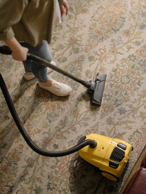 A professional cleaner from Croydon Carpet Cleaning is performing deep surface cleaning on a beige carpet inside a well-lit living room. The cleaner is dressed in full protective gear, including a white coverall, gloves, a face mask, and shoe covers, ensuring sanitisation and hygiene during the cleaning process. They are using a portable steam cleaning machine with a long wand to extract dirt and dust from the carpet fibers, which appears damp and in the process of being cleaned thoroughly. The room features natural lighting from a large window, with white walls, a wooden side table, and several potted plants contributing to a clean and inviting atmosphere. The process indicates a focus on deep cleaning and sanitisation, aligning with the services provided by Croydon Carpet Cleaning, aimed at maintaining healthy, spotless domestic surfaces.