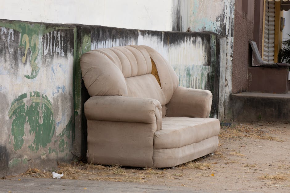 An old, beige upholstered armchair with a torn backrest sits outdoors against a weathered wall with peeling paint and faded graffiti. The armchair appears dusty and neglected, with dirt accumulation on the fabric and minor staining. It is positioned on a dirt ground with scattered dry leaves and debris nearby, suggesting outdoor exposure. In the background, there are steps leading up to a building with a partially open door, and a small plant is visible on the right side. This setting highlights the need for comprehensive cleaning and restoration, which Croydon Carpet Cleaning, as featured on croydoncarpetcleaning.org.uk, can provide through surface and deep cleaning services to restore such furniture to a hygienic condition.