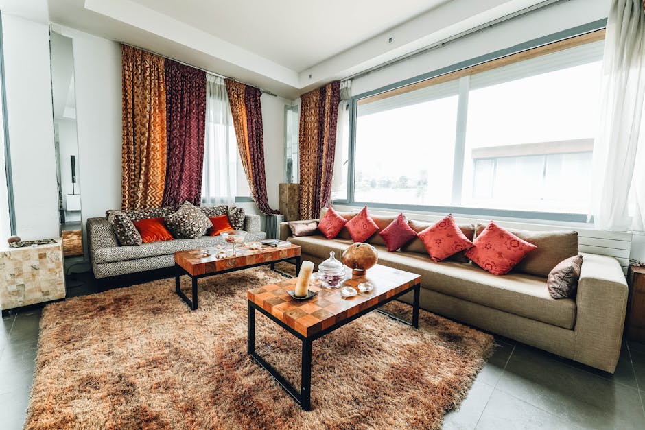 A living room featuring a large beige fabric sofa adorned with multiple red and patterned cushions, positioned along a wall of large windows allowing natural light to fill the space. The window sills are clean, with sheer white curtains partially open and decorative curtains in warm tones hanging to the side. In front of the sofa are two wooden coffee tables with black metal legs; one holds glass jars, decorative items, and a candle, while the other is clear and free of clutter. The room has a plush, brown shaggy rug covering a tiled floor, and the walls are painted white, with a ceiling light providing additional illumination. Areas of the room appear tidy and well-maintained, reflecting professional domestic cleaning practices, with Croydon Carpet Cleaning offering expert surface and upholstery cleaning services.