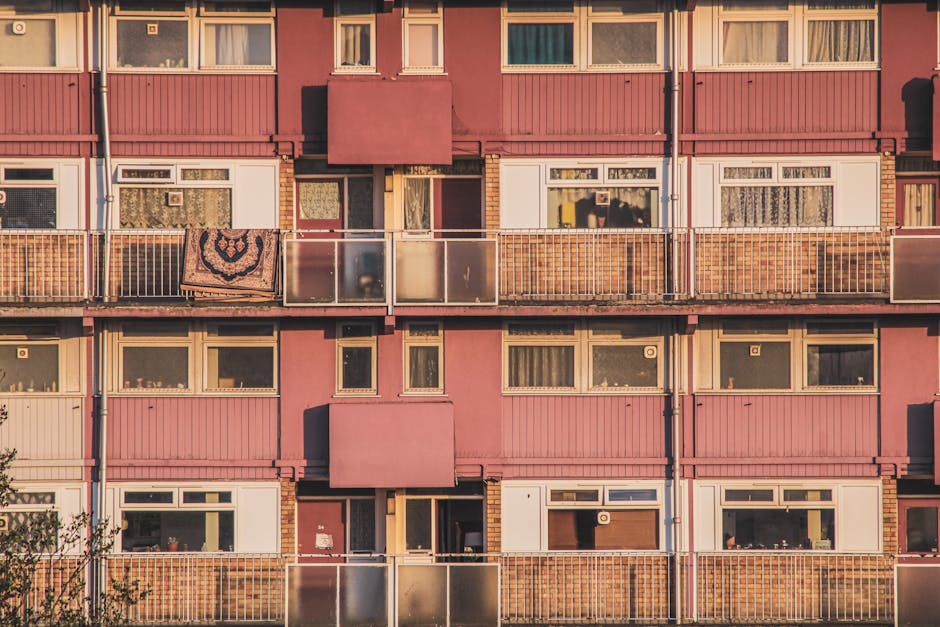 Exterior view of a pink residential apartment building with multiple floors, featuring balconies with metal railings and transparent glass panels. Some balconies have decorative items such as a patterned rug hanging over the railing. The windows vary in size, with some showing lace curtains and others revealing interior spaces. The building's facade includes brick and paneling sections, illuminated by warm sunlight, with shadows cast on the structure. This image, associated with Croydon Carpet Cleaning, emphasizes the importance of surface cleaning and maintenance in residential environments near East Croydon station.