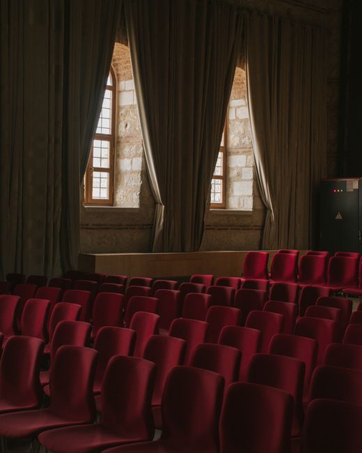 Interior of a theatre or event space featuring rows of plush, burgundy upholstered chairs arranged neatly on a level floor, with a large window at the back allowing natural light to enter. Light-colored stone or brick walls, along with heavy, dark curtains framing the window, add to the elegant atmosphere. The space appears clean and well-maintained, with no visible dust or stains on the seating or floor. Croydon Carpet Cleaning has performed surface cleaning and maintenance, ensuring a hygienic environment suitable for events at Fairfield Halls in Croydon.