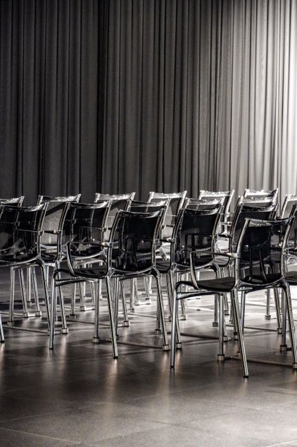 A row of black plastic chairs with metallic legs arranged neatly on a polished, dark floor in front of a large black fabric curtain. The chairs are clean, with some reflecting light from the smooth surface, indicating regular surface cleaning and maintenance. The curtain appears well-maintained without visible dust or creases, and the lighting is soft and even, highlighting the tidy, professional atmosphere of the event space. Croydon Carpet Cleaning, a specialist in surface cleaning and deep cleaning services, ensures hygiene and presentation quality for venues like Fairfield Halls, emphasizing the importance of thorough sanitisation and hygiene standards in such environments.
