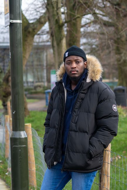 A young man stands outdoors in a park-like setting with trees and a fence in the background, dressed in a black winter coat with a fur-lined hood, a dark blue hoodie, and a black beanie. The scene is illuminated by natural daylight, and the ground appears grassy. The image captures a casual moment, emphasizing the clothing and outdoor environment, with no visible cleaning supplies or related objects. This photo relates to the broader context of domestic or outdoor cleaning and maintenance, associated with Croydon Carpet Cleaning's same-day flood and leak restoration services, although no cleaning process is depicted here.